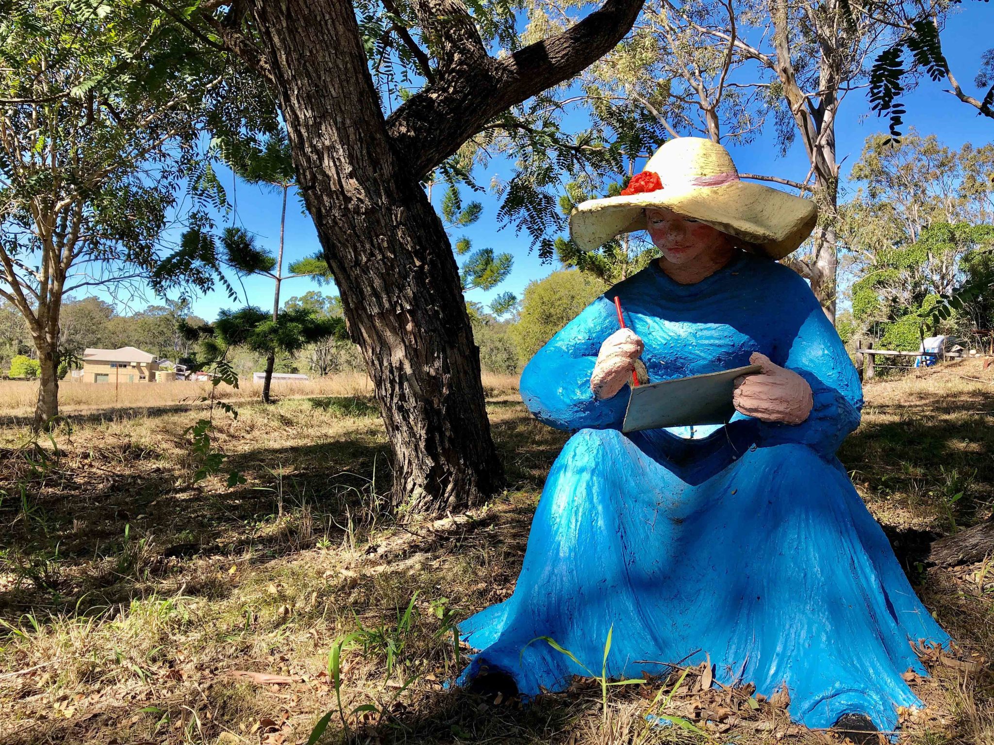 A “mystery woman” in a blue dress with pen and sketchpad in hand sits under a shady tree alongside the driveway into the property of Apple Tree Creek artist Alice McLaughlin.